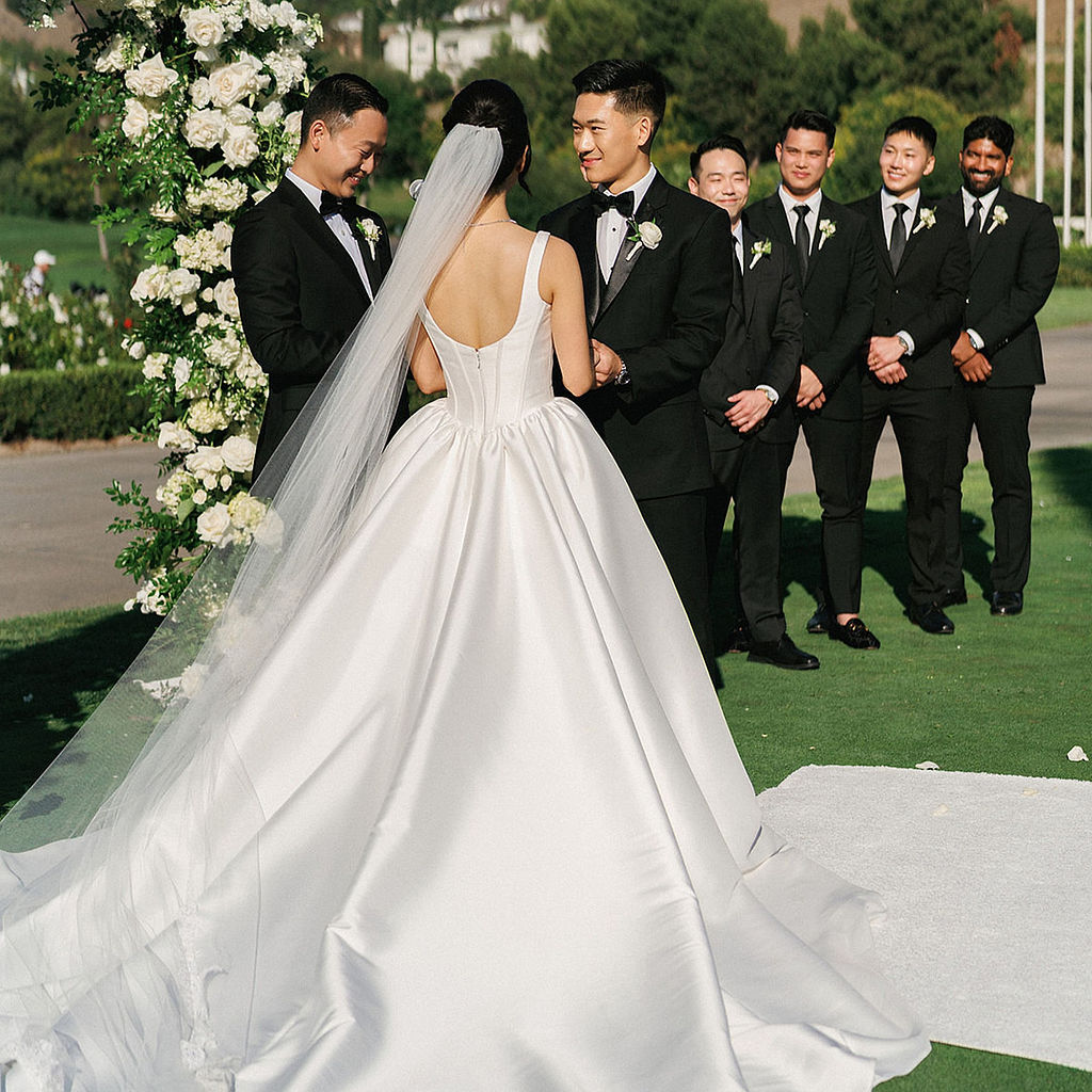 Bride in a satin ballgown facing her groom at the altar during a Marbella Country Club wedding ceremony, groomsmen in black tuxedos behind him