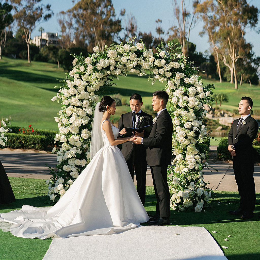 Bride in a satin ballgown facing her groom at the altar during a Marbella Country Club wedding ceremony, groomsmen in black tuxedos behind him