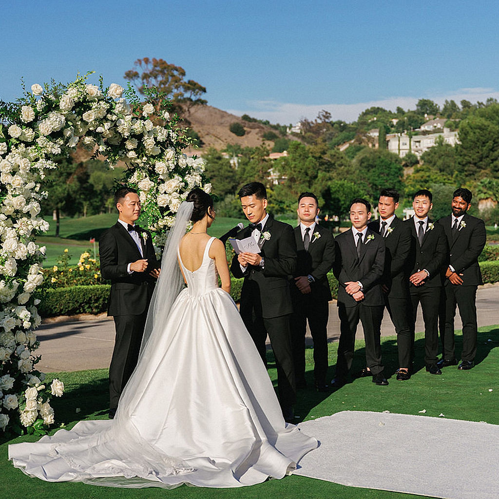 Bride in a satin ballgown facing her groom at the altar during a Marbella Country Club wedding ceremony, groomsmen in black tuxedos behind him