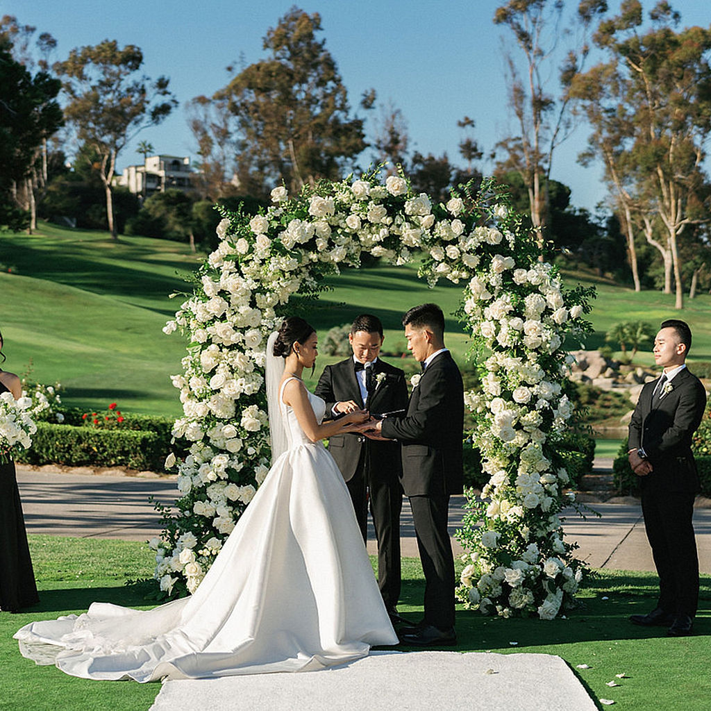 Bride and groom exchanging rings under a white rose arch on the golf course at Marbella Country Club in San Juan Capistrano