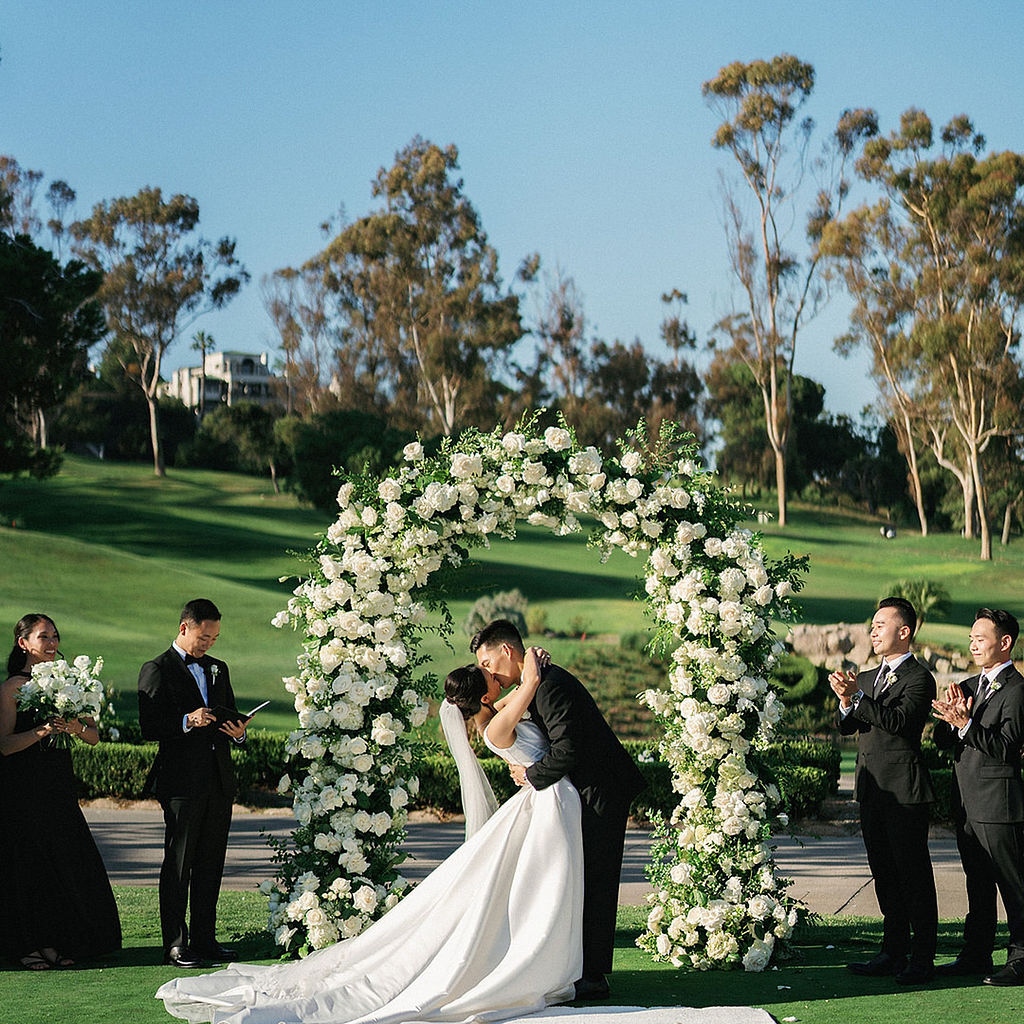 Groom dipping his bride for their first kiss under a circular white rose and greenery arch, wedding party applauding