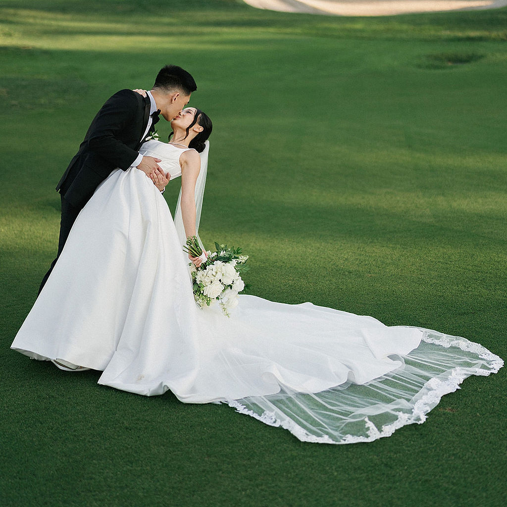 Bride and groom kissing on the fairway at golden hour, her lace-edged cathedral veil spread across the grass
