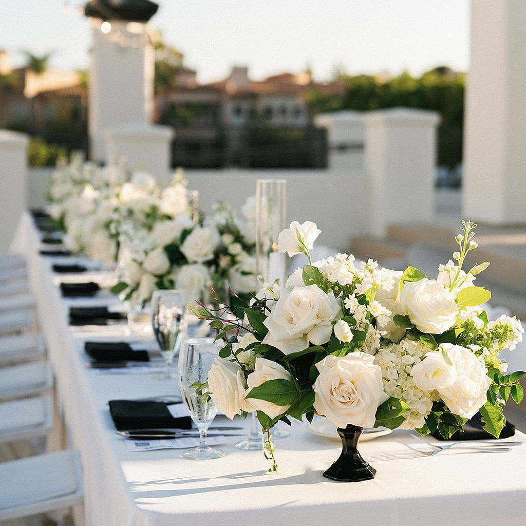 Long reception table with white linens, black napkins, and low white rose centerpieces in black compotes