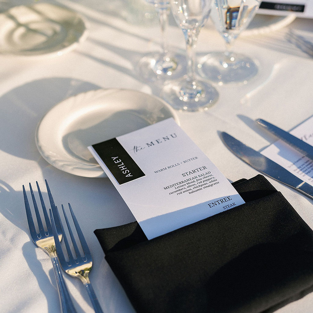 Black and white printed menu card tucked into a folded black napkin at a wedding place setting