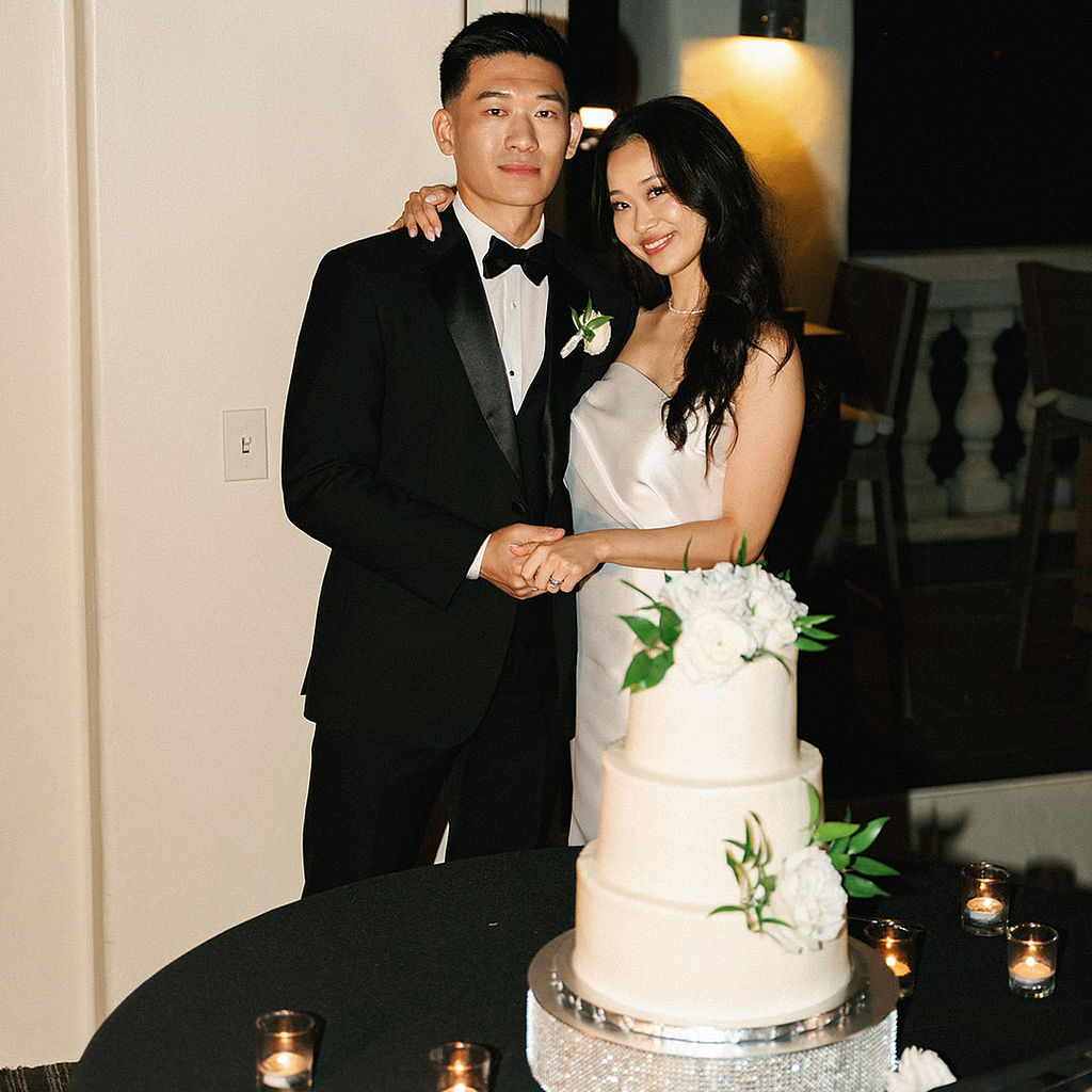 Bride and groom holding hands beside their three-tier white wedding cake trimmed with fresh roses