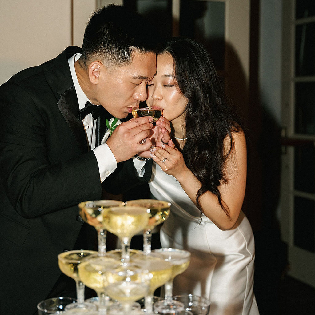 Bride and groom drinking from the top coupes of a champagne tower at their reception